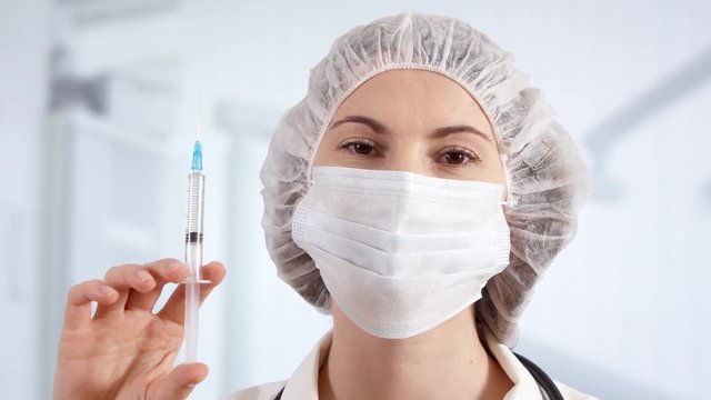 Professional female doctor in mask, cap and gloves in hospital room with disposable one-off syringe with transparent drug. Woman physician at work. Laboratory employee making scientific research