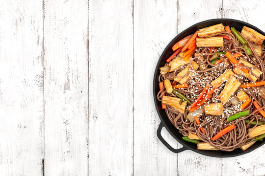 Delicious soba with tofu skin and vegetables in a cast iron pan on light wooden background, top view