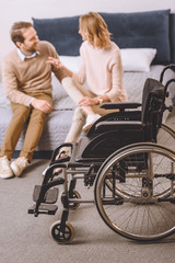 husband with disability and wife sitting and talking on bed with wheelchair on foreground