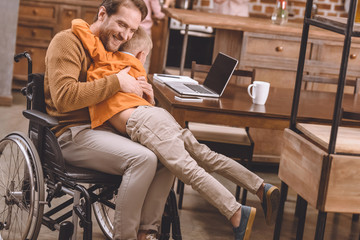 happy disabled father in wheelchair hugging with cute little son at home