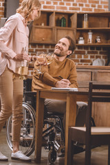 cropped shot of smiling woman holding wine glasses and bottle while husband in wheelchair taking notes at home