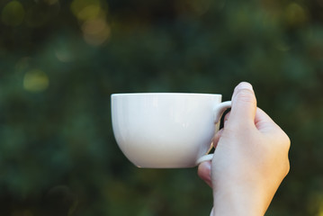 Woman hand holding a cup of coffee in the morning, with nature background. Relaxing and healthy concept.