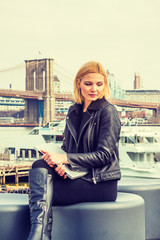 Fototapeta na wymiar Businesswoman traveling, working in New York, wearing black leather jacket, sitting by river, holding laptop computer, looking down, sad, thinking. Brooklyn Bridge, boat on background. Filtered effect