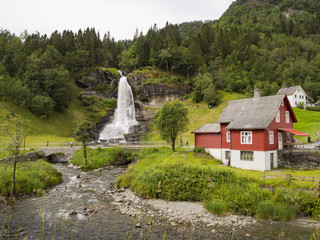 Fototapeta na wymiar Paisaje alpino en Norheimsund, norte del Hardangerfjord, donde se ubica la impresionante cascada Steinsdalsfossen, rodeada de vegetación y una casita roja típica Noruega, en el verano de 2017