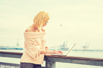 Fototapeta na wymiar Eastern European Businesswoman traveling, working in New York, wearing off shoulder sweater, glasses, working on laptop computer by river in sunny spring day, looking down, typing. Filtered effect..