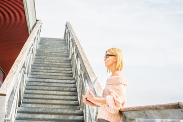 Way to Success. European college student studying in New York, wearing off shoulder knitted sweater, glasses, working on laptop computer by stairs on campus, looking up, thinking. Filtered effect.