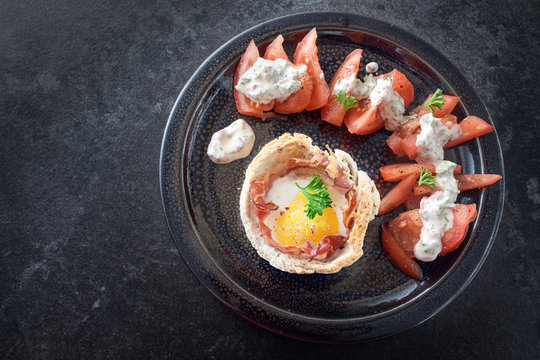 egg muffin with bacon in toasted bread, tomato with cheese cream and parsley garnish on a black plate on a dark slate background with copy space, high angle top view from above