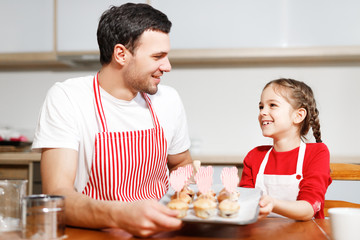 Obraz na płótnie Canvas Indoor shot of handsome man wears striped apron and his small pretty female kid hold hand made cakes, rejoices successfully done work on kitchen, being in high spirit, happy spend time together.