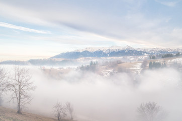Winter landscape in the mountains. Rural landscape in the evening with fog and clouds on the sky. There are some houses in the image.