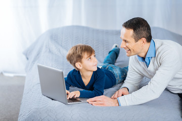 Fondly look. Happy young man lying on the bed next to his little son and smiling at him while the boy using laptop and smiling back at his father