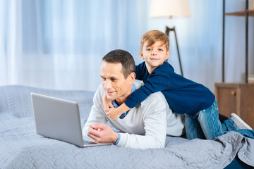 Gentle hug. Adorable little boy back-hugging his beloved father lying on the bed and working on the laptop