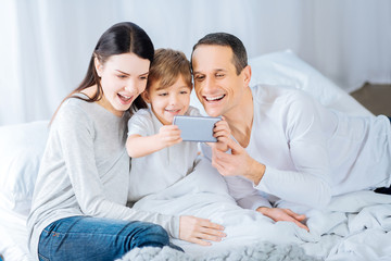 Sweet moments. Joyful little boy sitting in the bed under the blanket and using a phone to take a selfie with his beloved parents