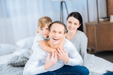 Happiest day. Upbeat young man sitting on the bed and posing for family portrait together with his son back-hugging him and his pretty wife sitting slightly behind him