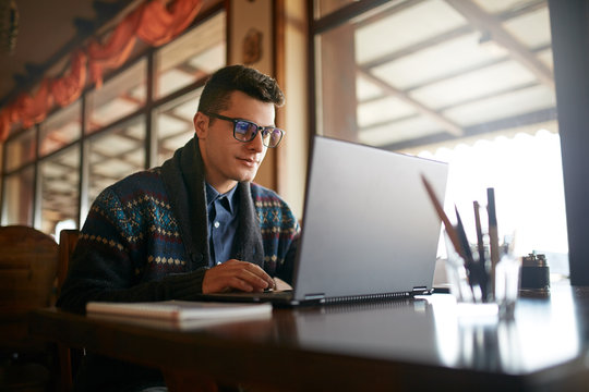 Handsome freelancer businessman working on laptop in cafe. Blogger man updating his profile in social networks with photos sharing with followers multimedia using notebook with wifi. Nerd in glasses.