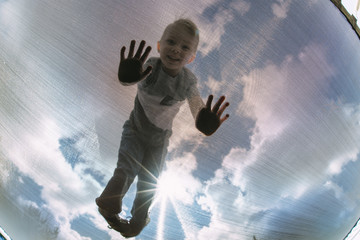 Low angle portrait of boy playing on trampoline against cloudy sky seen through net