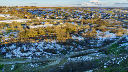 Fototapeta na wymiar Top view aerial rural countryside winter or early spring landscape. Dirt road, village houses and riever. Fields covered by young green grass and snow. Copy space long image background.