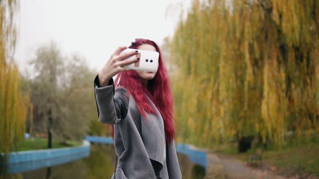 Young attractive woman with red hait making selfie on her smartphone while standing by an artificial pond in an autumn park