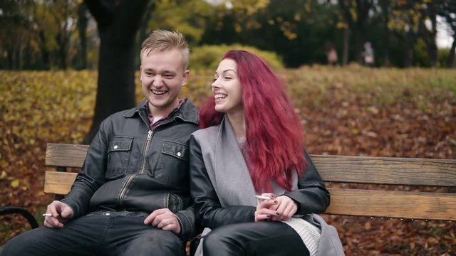 Young attractive couple sitting on a bench in autumn park and talking. Attractive woman with red hair is smoking