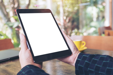 Mockup image of hands holding black tablet pc with blank desktop white screen and coffee cup on wooden table in cafe background