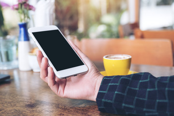Mockup image of a hand holding white mobile phone with blank black desktop screen and yellow coffee cup on wooden table in modern cafe