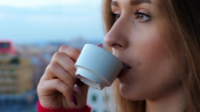 Beautiful young red-haired student female standing on the terrace in the centre of mega polis. Pretty girl dressed in red sweater taking a cup of tasty coffee and watching city hustle and bustle