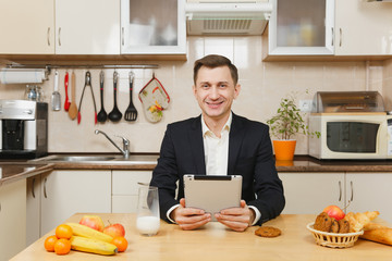 Handsome European young business man in suit, shirt having breakfast at home, sitting at table with tablet, fruits, eating oat cookies with milk on light kitchen. Dieting concept. Healthy lifestyle.