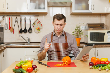 Handsome caucasian young man in an apron, brown shirt sitting at table, looking recipe in tablet, cuting vegetable for salad with knife in light kitchen. Dieting concept. Cooking at home. Prepare food