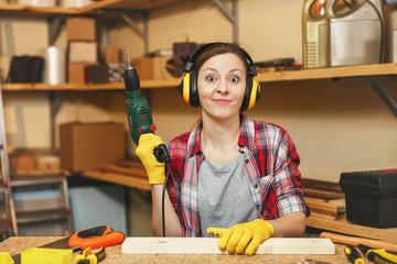 Beautiful caucasian young brown-hair woman in plaid shirt, noise insulated headphones working in carpentry workshop at table place, drilling with power drill holes in piece of wood, making furniture.