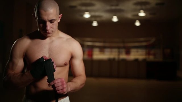 A pumped-up man in boxing boots and shorts in the gym against the background of a boxing ring dresses bandages on his hands.