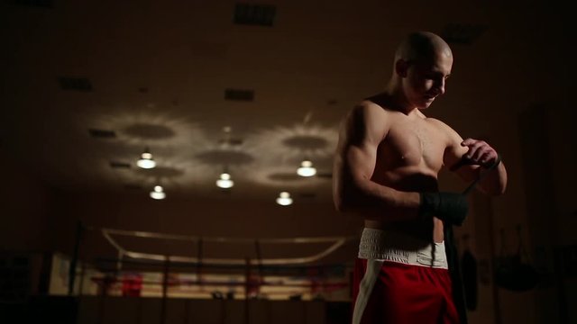 A pumped-up man in boxing boots and shorts in the gym against the background of a boxing ring dresses bandages on his hands.