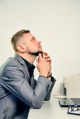 man with chin in hands thoughtfully looking upwards sitting at table with laptop in office