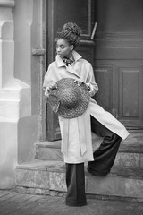African girl on the street in a blue coat holding a Burgundy hat with two hands against street stairs and the blue door black and white image