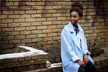 African girl in the blue coat in the style fashion sitting on the ledge and background the yellow brick wall