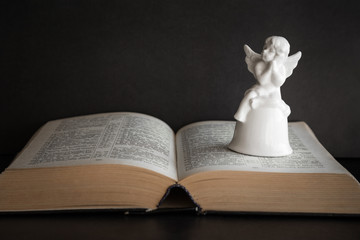 Little, white angel figurine on the opened bible. Quiet, dark atmosphere. Prayer time on sunday.