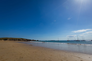 Wide open sandy beach at low tide with sailboats moored offshore and distant people walking at the edge of the gentle surf