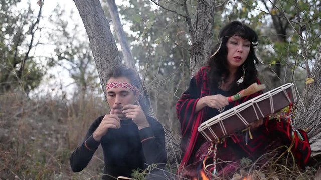 shamanic woman playing on shaman frame drum in the nature around the fire and a man playing a harp