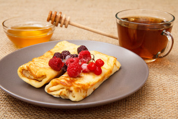 Homemade pancakes filled with cottage cheese and topped with frozen raspberries and blackberries on plate, cup of tea and honey in glass bowl with wooden spoon