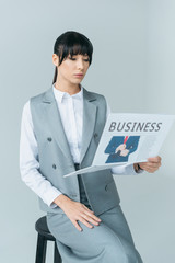 businesswoman sitting on chair and reading business newspaper isolated on gray