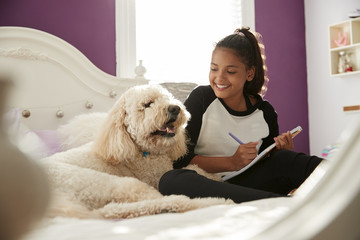 Young teen girl doing homework on her bed with pet dog - Powered by Adobe