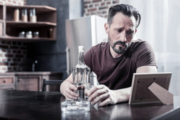 My memories. Sad gloomy cheerless man sitting at the table and looking at the photo while drinking alcohol