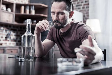 Chronic depression. Serious thoughtful cheerless man sitting at home and drinking vodka while focusing on his problems