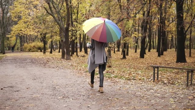 Back view of a young woman with red hair walking in autumn park with colorful rainbow umbrella and holding paper cup with coffee. Girl in warm coat enjoying cool fall weather with a cup of hot drink