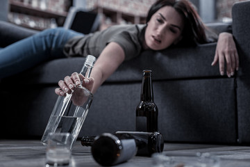 Not enough. Selective focus of a bottle with alcohol being taken by a drunk unhappy woman while lying on the sofa