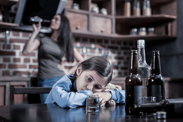 Fototapeta na wymiar Alcohol addicted parents. Cheerless unhappy cute girl sitting at the table and being thoughtful while looking at the beer bottles