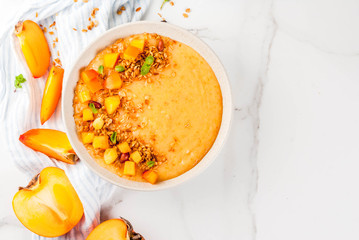 Diet healthy vegan food recipes, delicious persimmon smoothie bowl with smooth and creamy persimmon and crunchy spiced granola, white marble table, top view copy space