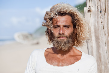 Robinson Crusoe. Portrait of curly-bearded man on the beah with a ship wreckage on the background.