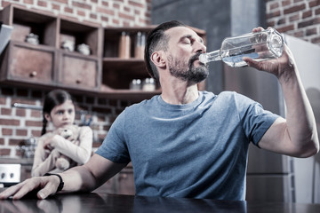 Naklejka na ściany i meble Need for the bottle. Sad depressed drunk man sitting in the kitchen and holding a bottle of vodka while drinking