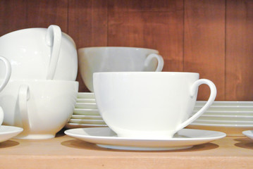 Bright white dishes, plates and cups standing on brown wooden shelf. Concept of buying choosing new dishes for house home, interior indoor decoration o for gifts. Clean ordered dishes in store.