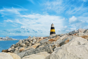 Little beautiful white, yellow and black lighthouse standing over the big stones at the pier of Benalmadena port, panoramic view to a seashore with hotels, resorts and beach. Spain winter vacation.