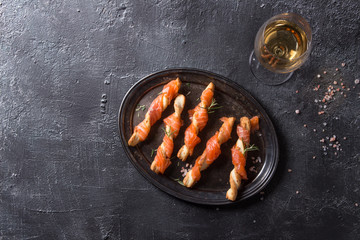 Appetizer with smoked salmon on crispy breadsticks served on vintage metal tray with sea salt, glass of white wine over black texture background. Top view with space